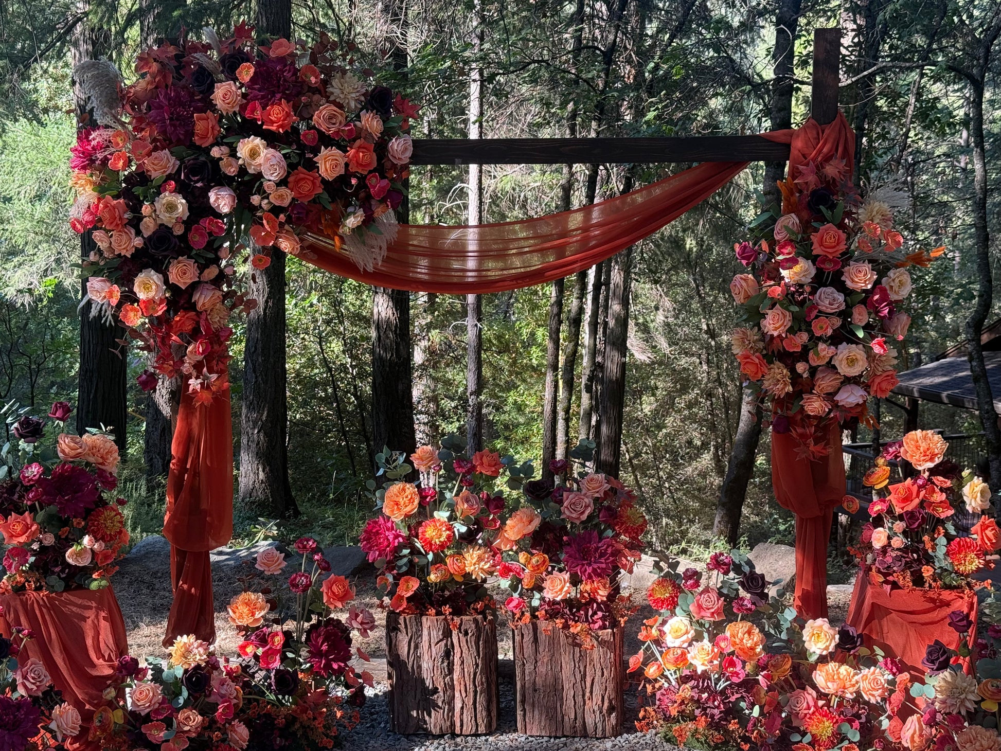 Full Evermore Fall ceremony setup with arch adornment rental, aisle flower rentals, and rust chiffon drapery with coral, peach, and burgundy florals and wood pedestal wraps at outdoor redwood forest wedding | Reno, Lake Tahoe & Sacramento