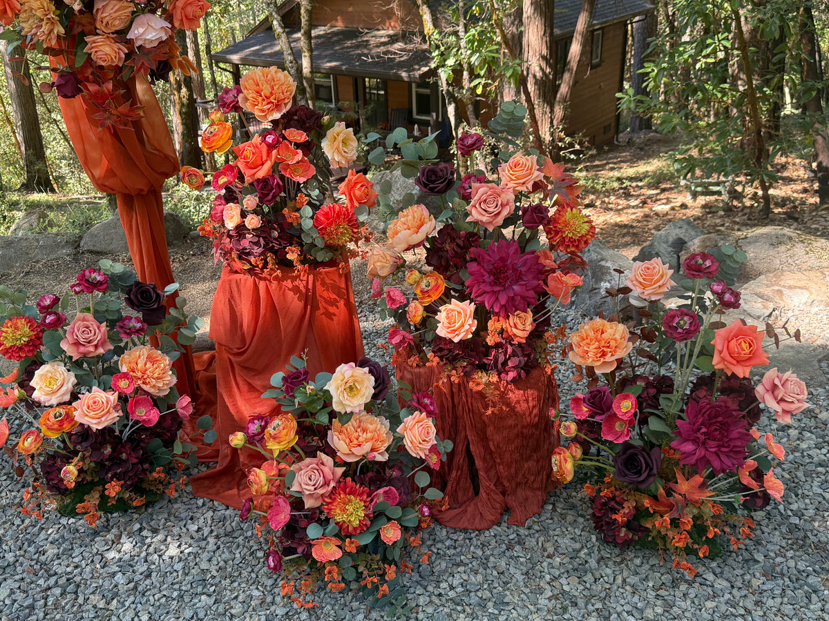 Evermore Fall aisle flower rentals with coral roses, orange ranunculus, burgundy dahlias, and rust chiffon drapery beside Evermore arch adornment at outdoor redwood forest wedding | Reno, Lake Tahoe & Sacramento
