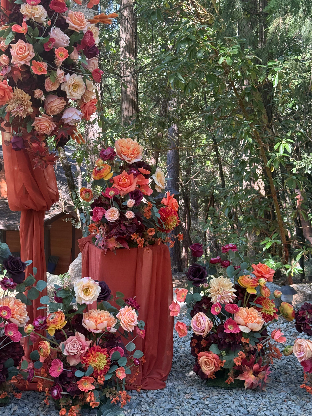 Evermore Fall aisle flower rentals with coral roses, orange ranunculus, burgundy dahlias, and rust chiffon drapery on 24" pedestals and at the base of Evermore arch adornment at outdoor redwood forest wedding | Reno, Lake Tahoe & Sacramento
