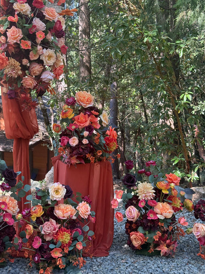 Evermore Fall aisle flower rentals with coral roses, orange ranunculus, burgundy dahlias, and rust chiffon drapery on 24" pedestals and at the base of Evermore arch adornment at outdoor redwood forest wedding | Reno, Lake Tahoe & Sacramento