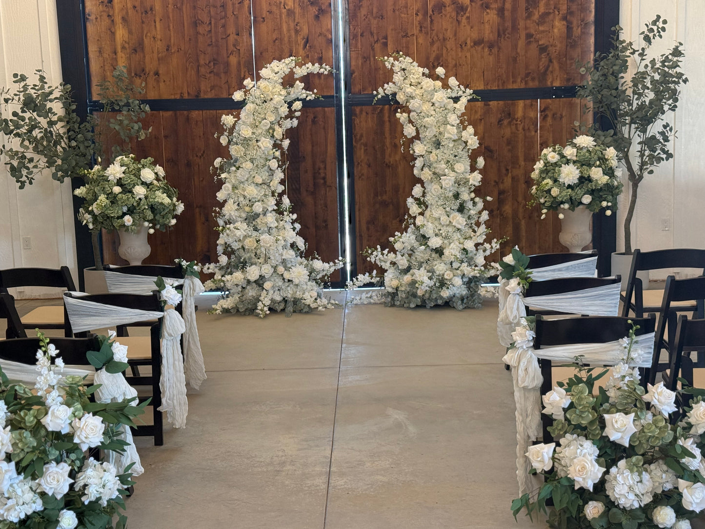 Fairytale double moon arch rentals with white roses and lush greenery at indoor barn wedding ceremony with Enchanted aisle flowers, black folding chairs with white ties, and faux eucalyptus trees in barn setting | Reno, Lake Tahoe & Sacramento
