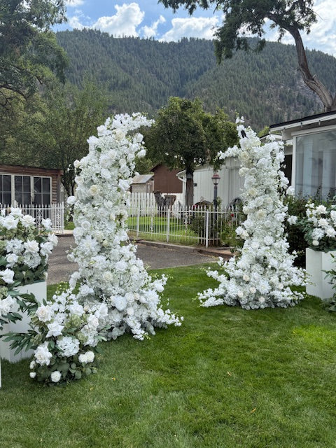Full setup of Fairytale double moon arch rentals with Enchanted aisle flowers with white roses and lush greenery on grass with white pedestal boxes at base at outdoor mountain wedding venue | Reno, Lake Tahoe & Sacramento