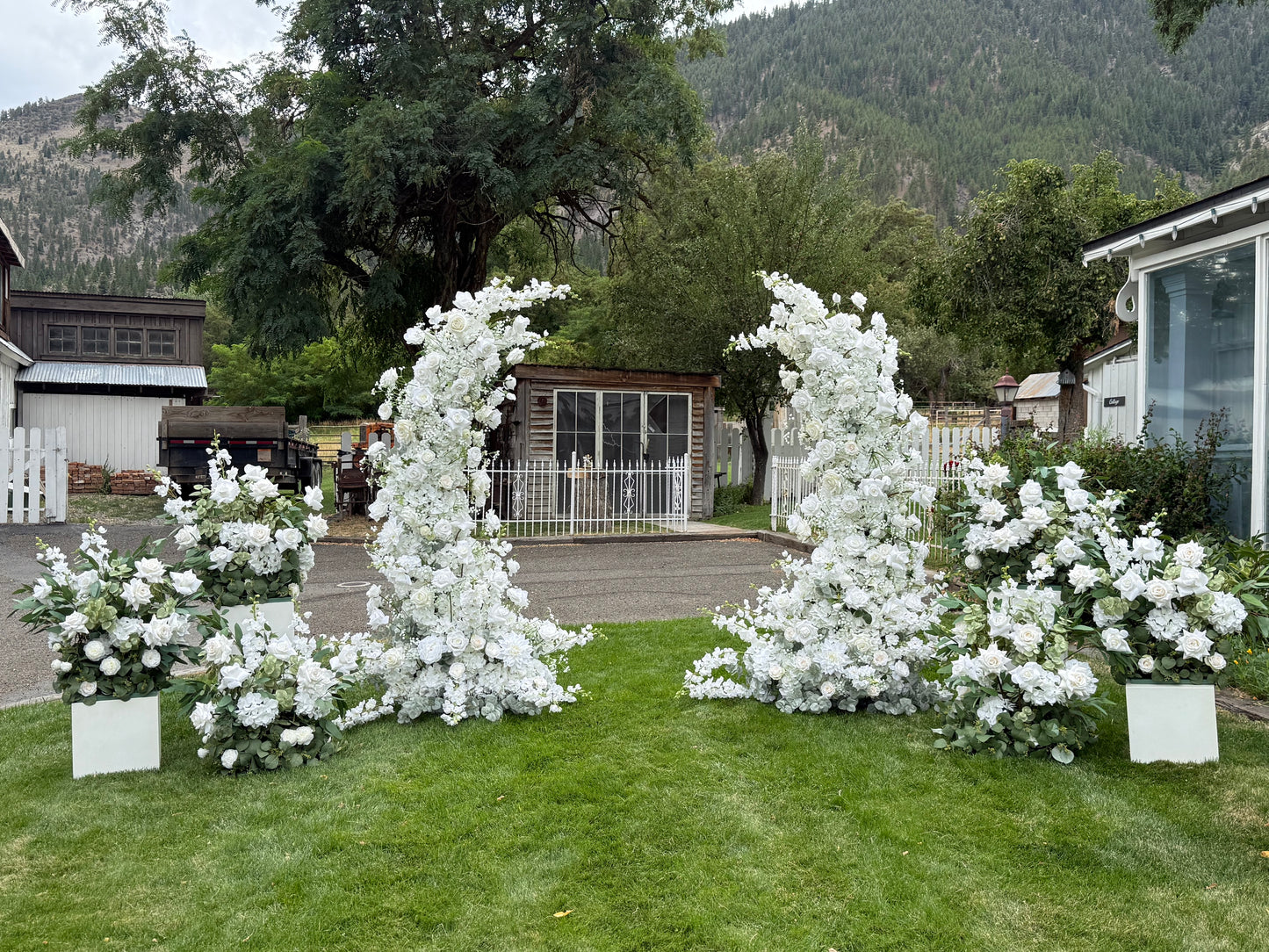 Wide setup view of Fairytale double moon arch rentals with white roses and lush greenery and four white pedestal boxes with the Enchanted aisle flowers on them at base at outdoor mountain wedding venue | Reno, Lake Tahoe & Sacramento