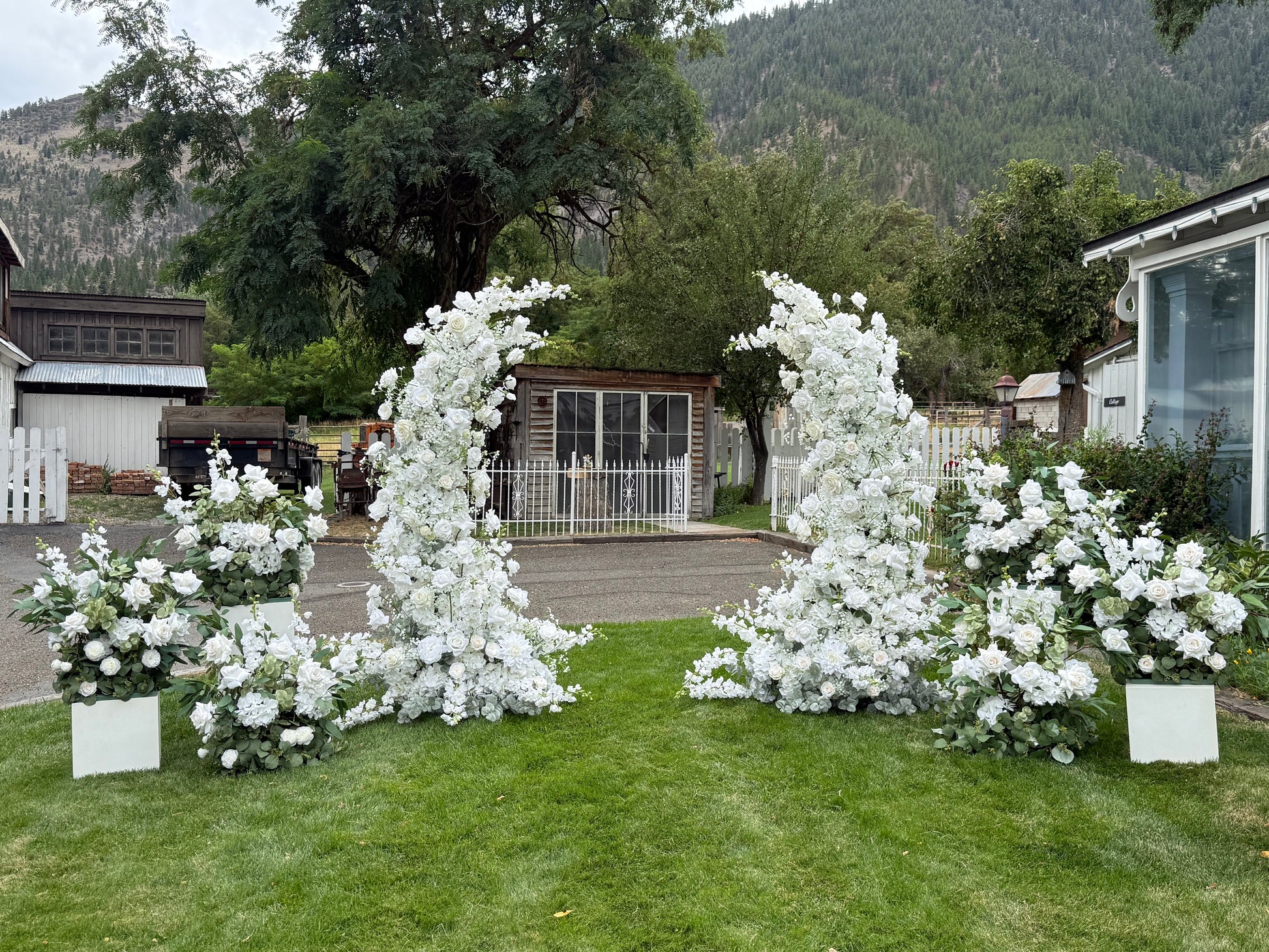 Wide setup view of Fairytale double moon arch rentals with white roses and lush greenery and four white pedestal boxes with the Enchanted aisle flowers on them at base at outdoor mountain wedding venue | Reno, Lake Tahoe & Sacramento