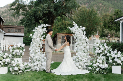 Bride and groom holding hands between Fairytale double moon arch rentals with white roses and lush greenery at outdoor mountain wedding venue with white pedestal boxes at base | Reno, Lake Tahoe & Sacramento