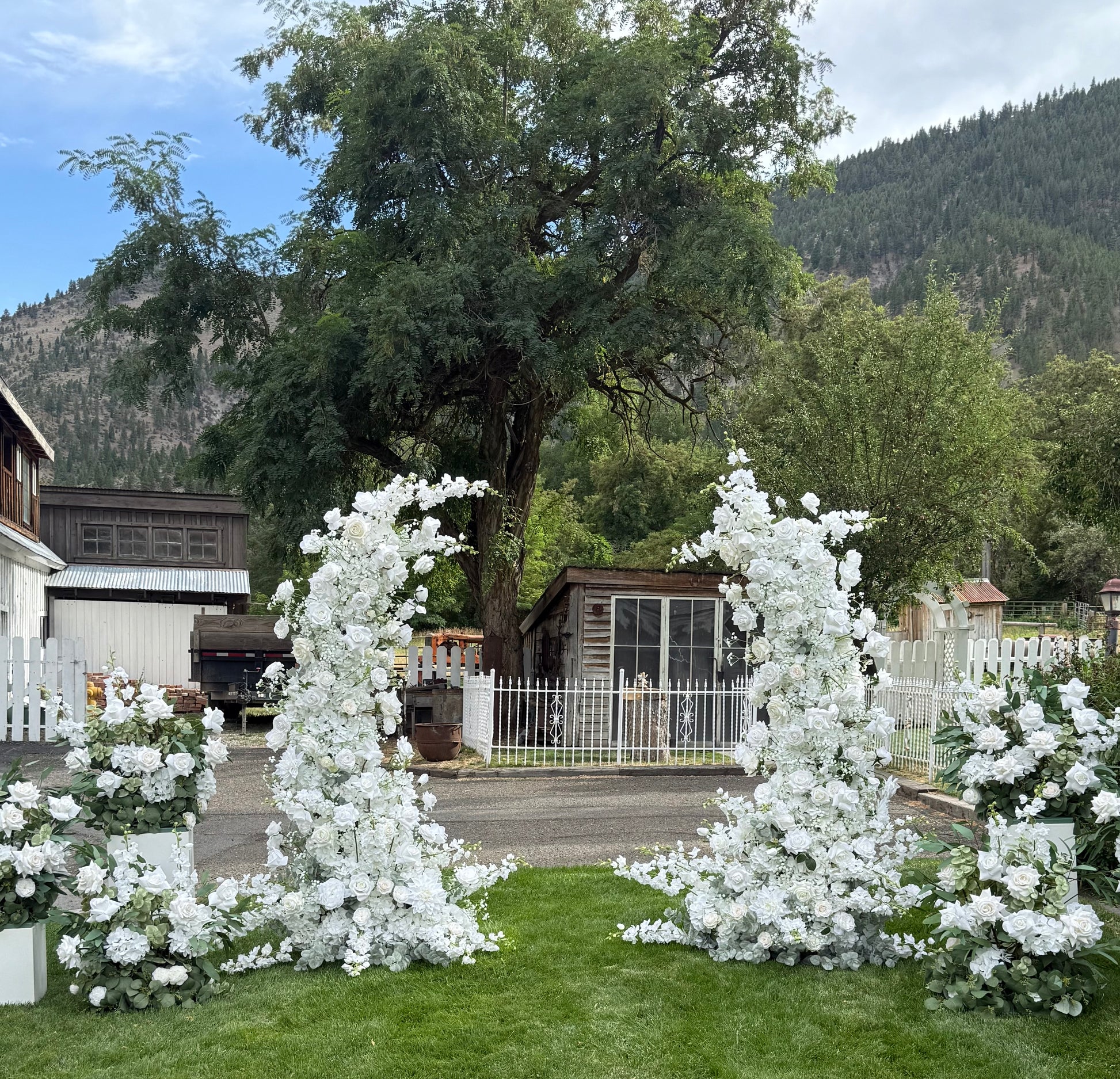 Wide view of Fairytale double moon arch rentals with white roses and lush greenery on grass with white pedestal boxes at base at outdoor mountain wedding venue | Reno, Lake Tahoe & Sacramento