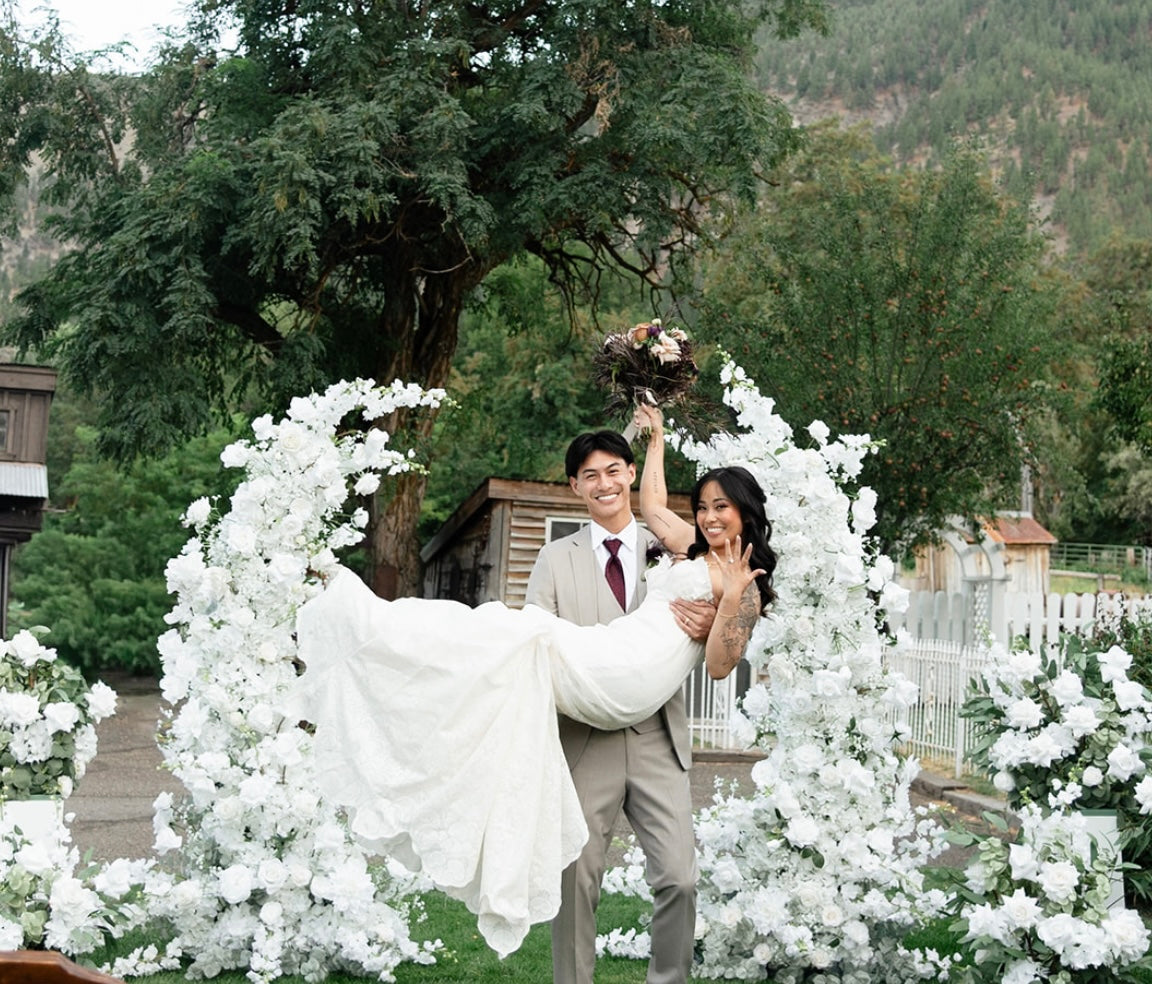 Groom carrying bride in front of Fairytale moon arch rental with white roses and lush greenery at outdoor mountain wedding venue | Reno, Lake Tahoe & Sacramento