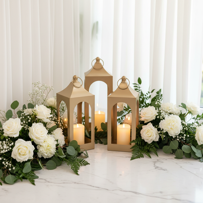Golden Lantern Rentals set of 3 with glowing flameless pillar candles arranged as elegant wedding corner vignette on white marble floor with lush white floral arrangements, greenery, and white drapery in background | Reno, Lake Tahoe & Sacramento