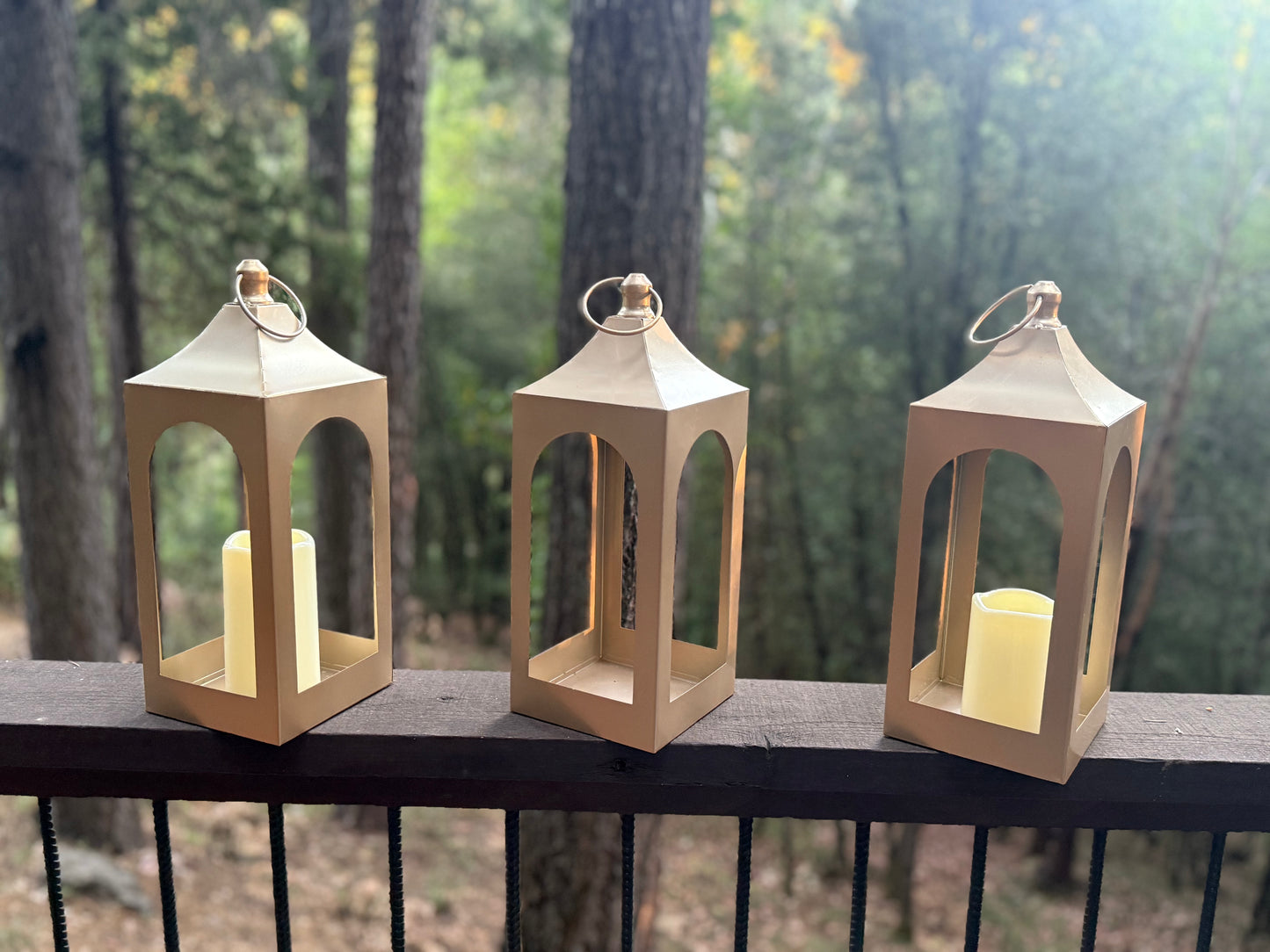 Golden Lantern Rentals set of 3 with flameless pillar candles displayed on dark wood railing with lush forest backdrop in natural daylight | Reno, Lake Tahoe & Sacramento