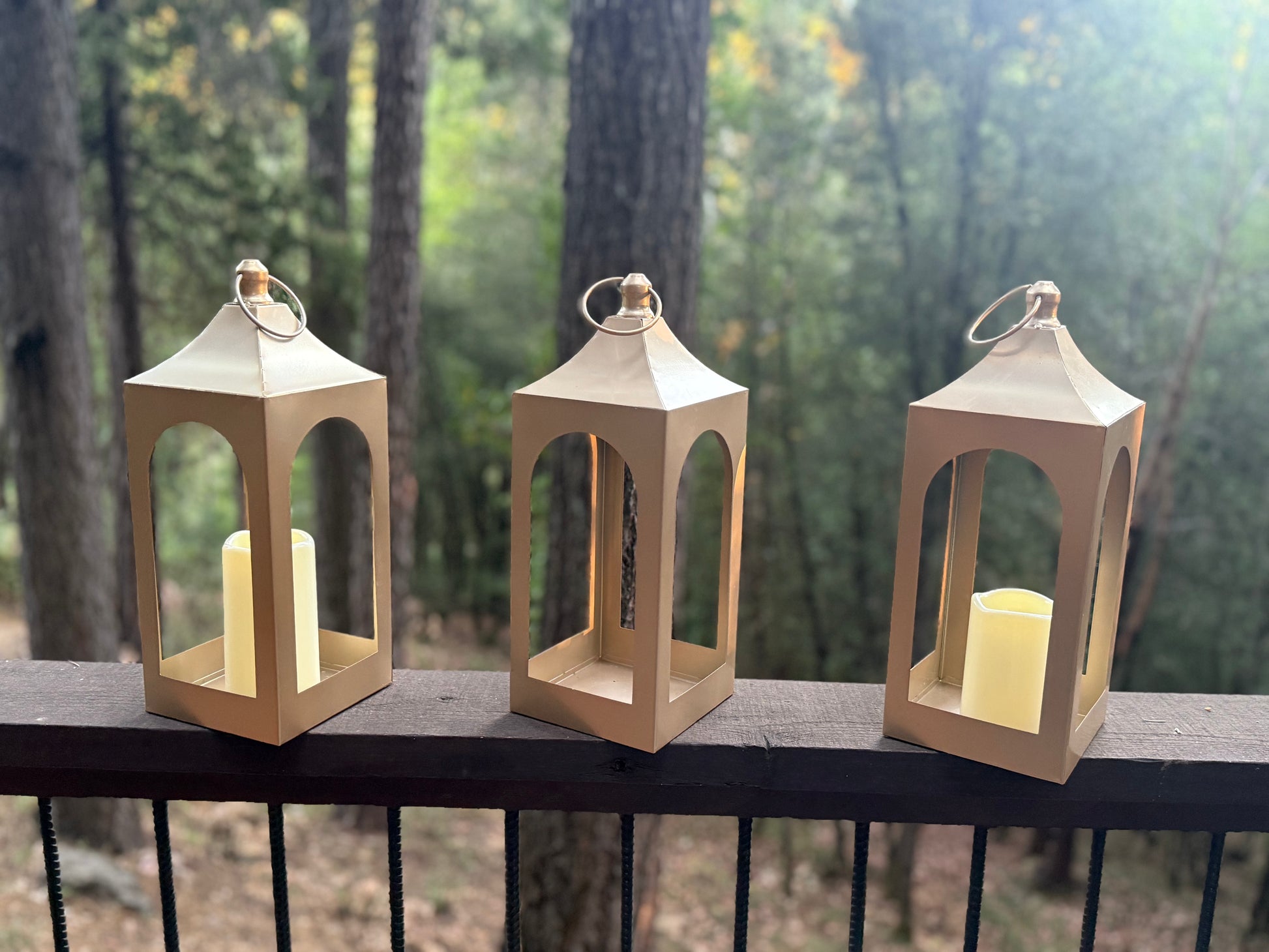 Golden Lantern Rentals set of 3 with flameless pillar candles displayed on dark wood railing with lush forest backdrop in natural daylight | Reno, Lake Tahoe & Sacramento