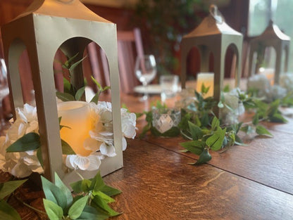 Golden Lantern Rentals set of 3 with glowing flameless pillar candles and white roses on rustic wood reception table with greenery garland, warm candlelit ambiance | Reno, Lake Tahoe & Sacramento