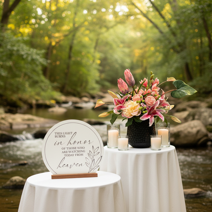 In Memory Sign Rental with Candle on white linen side table at outdoor wedding beside Tropical Blush large floral centerpiece of pink stargazer lilies, blush protea, and peach garden roses in textured black vase with white pillar candles and gentle creek and lush greenery in background | Reno, Lake Tahoe & Sacramento