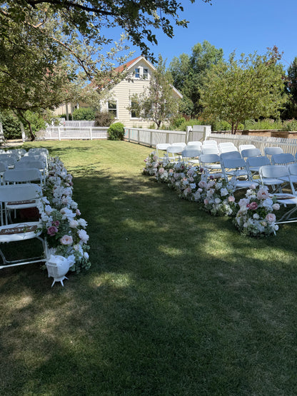 Wide view of Love Story aisle flower rentals lining both sides of outdoor grass ceremony aisle with blush, mauve, and white roses and white folding chairs at garden estate wedding venue | Reno, Lake Tahoe & Sacramento