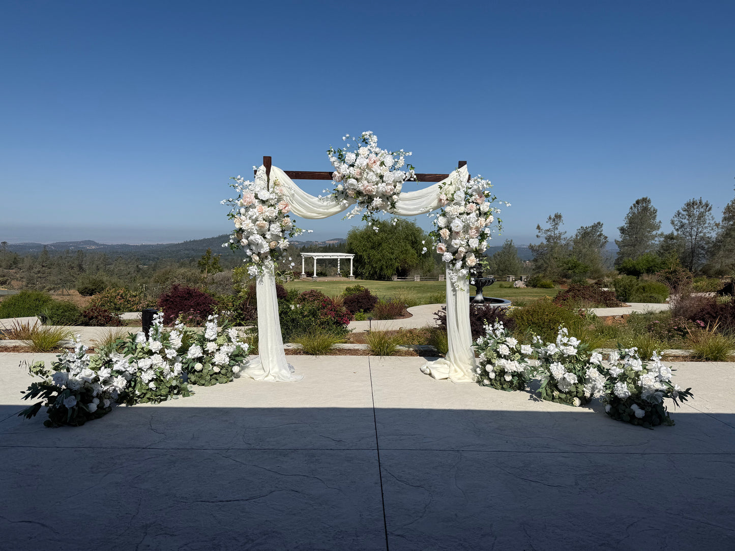 Full setup of Love Story arch adornment rental with blush and white roses, white drapery, and Enchanted aisle flower rentals flanking rustic wood arch at outdoor hilltop wedding venue with panoramic views | Reno, Lake Tahoe & Sacramento