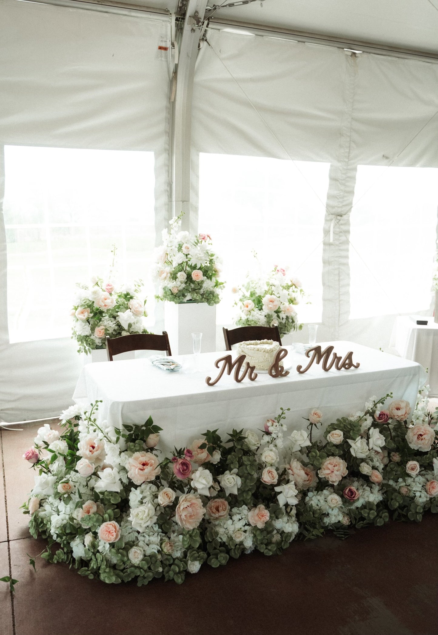 Mr & Mrs Wood Sign Rental on sweetheart table with Love Story aisle flower in lush blush and white floral table skirt of garden roses, hydrangea, and eucalyptus with tall white pedestals with more aisle flowers in elegant white tent wedding reception | Reno, Lake Tahoe & Sacramento 