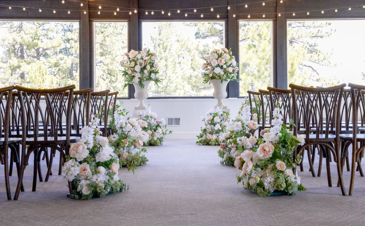 Two Love Story floral stone urn rentals on white pedestals flanking indoor wedding ceremony altar with Love Story aisle flowers in blush and white roses, cross-back chairs, and string lights at Lake Tahoe venue | Reno, Lake Tahoe & Sacramento