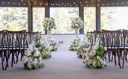 Two Love Story floral stone urn rentals on white pedestals flanking indoor wedding ceremony altar with Love Story aisle flowers in blush and white roses, cross-back chairs, and string lights at Lake Tahoe venue | Reno, Lake Tahoe & Sacramento