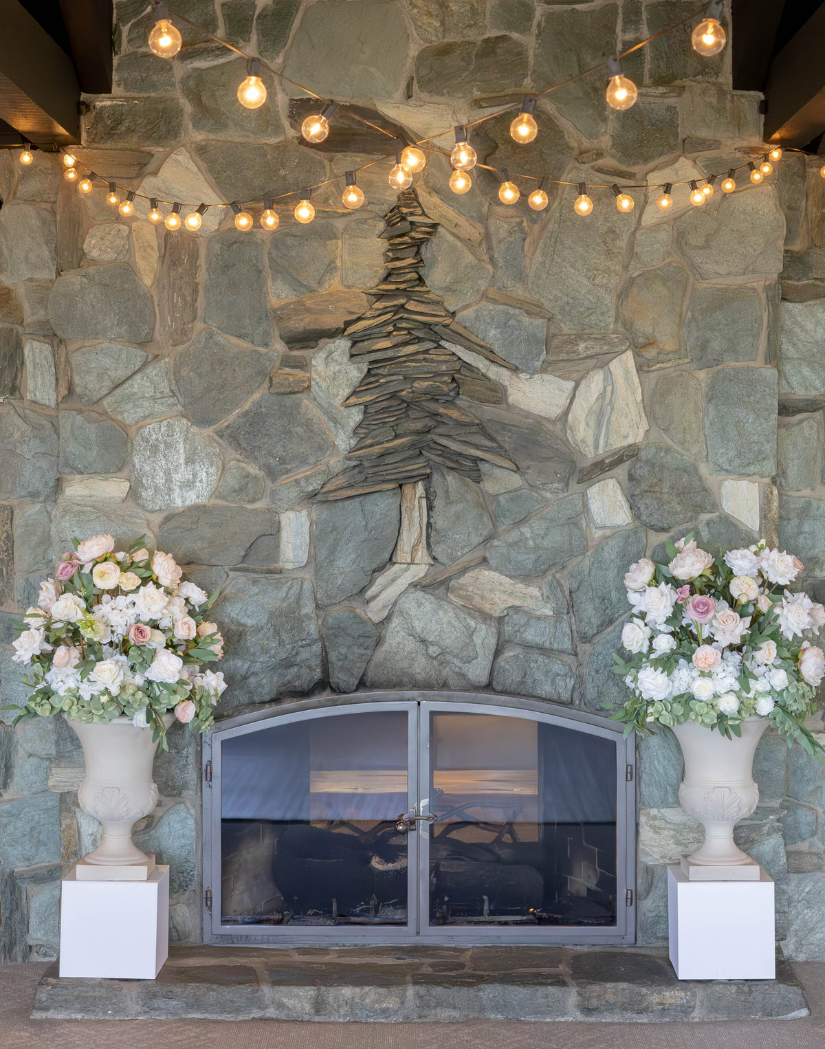 Two Love Story floral stone urn rentals on white square pedestals flanking stone fireplace with string lights and blush and white roses at indoor Lake Tahoe wedding venue | Reno, Lake Tahoe & Sacramento