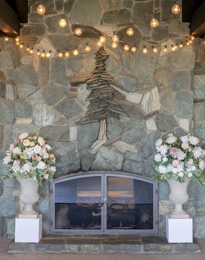 Two Love Story floral stone urn rentals on white square pedestals flanking stone fireplace with string lights and blush and white roses at indoor Lake Tahoe wedding venue | Reno, Lake Tahoe & Sacramento