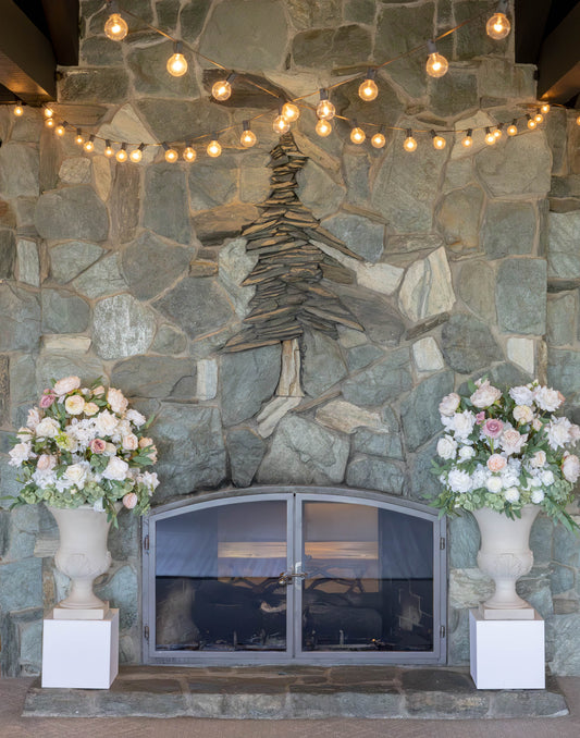 Two Love Story floral stone urn rentals on white square pedestals flanking stone fireplace with string lights and blush and white roses at indoor Lake Tahoe wedding venue | Reno, Lake Tahoe & Sacramento