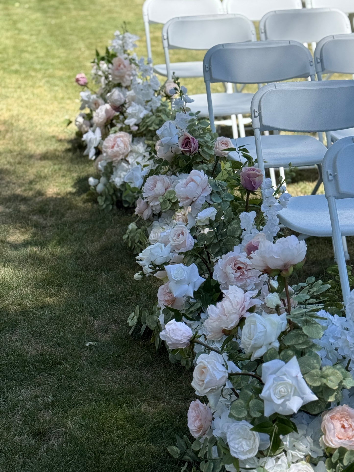 Side view of Love Story aisle flower rental row with blush, mauve, and white garden roses, peonies, and eucalyptus lining outdoor grass ceremony aisle beside white folding chairs | Reno, Lake Tahoe & Sacramento