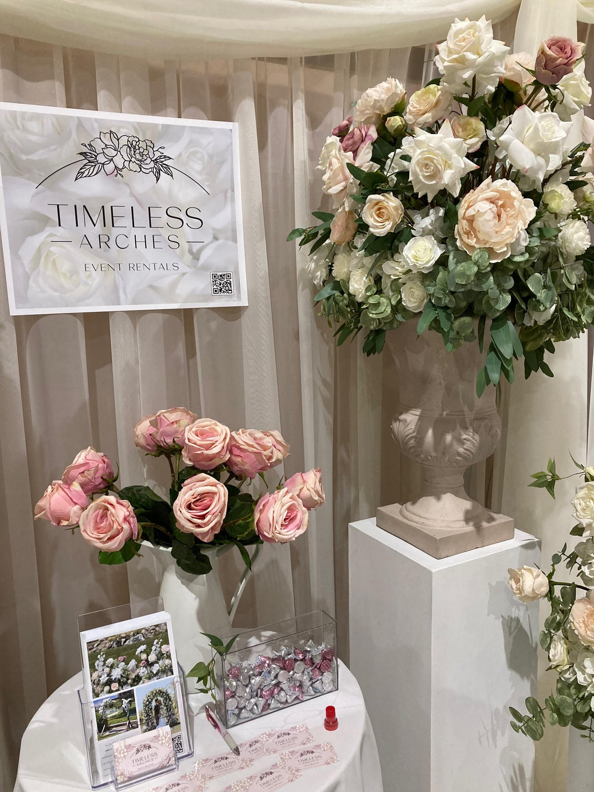 Timeless Arches event rental booth displaying Love Story floral stone urn on white pedestal with blush and cream roses and peonies at wedding bridal expo | Reno, Lake Tahoe & Sacramento