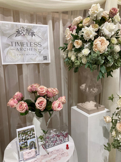 Timeless Arches event rental booth displaying Love Story floral stone urn on white pedestal with blush and cream roses and peonies at wedding bridal expo | Reno, Lake Tahoe & Sacramento