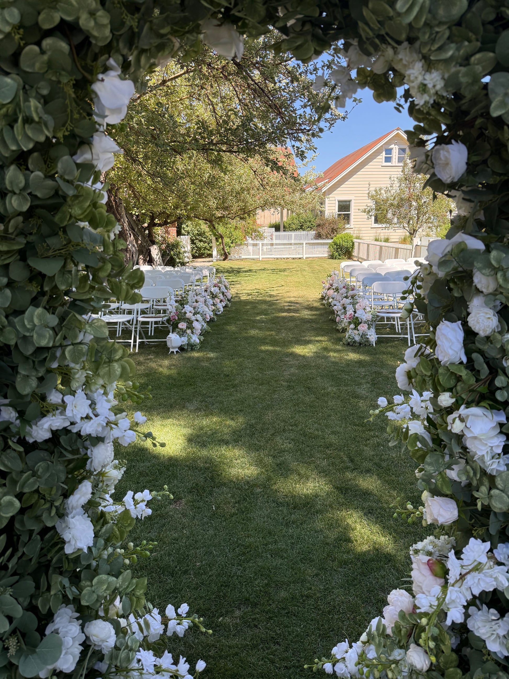 Love Story aisle flower rentals lining outdoor grass ceremony aisle viewed through floral arch with white roses and eucalyptus, white folding chairs on both sides | Reno, Lake Tahoe & Sacramento