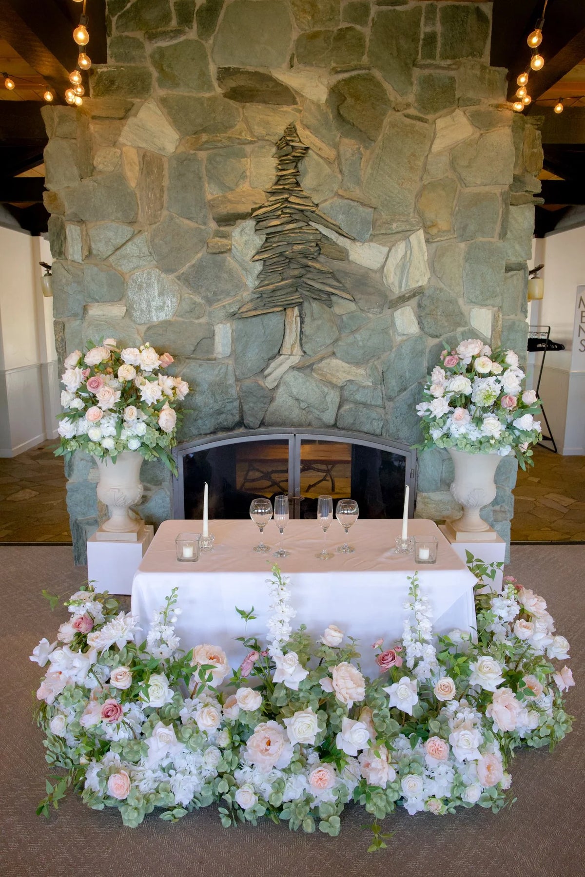 Love Story aisle flower and urn rentals styled as sweetheart table with white and blush roses in front of stone fireplace at indoor wedding venue | Reno, Lake Tahoe & Sacramento