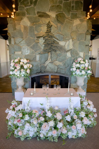 Love Story aisle flower and urn rentals styled as sweetheart table with white and blush roses in front of stone fireplace at indoor wedding venue | Reno, Lake Tahoe & Sacramento