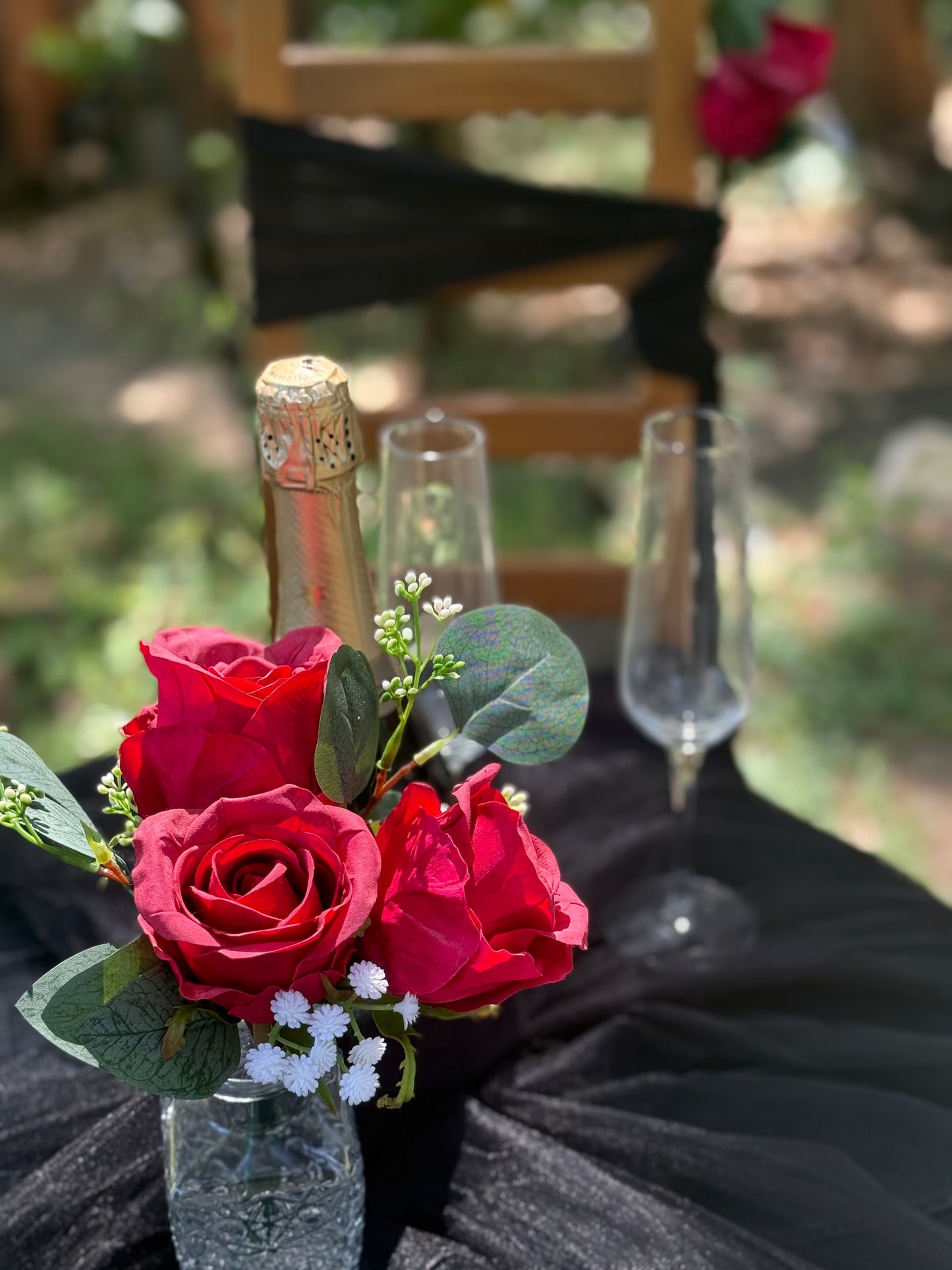 Red rose centerpiece rental with bold red roses, baby's breath, and eucalyptus in crystal bud vase styled on black linen reception table with champagne bottle and flutes at outdoor forest wedding | Reno, Lake Tahoe & Sacramento