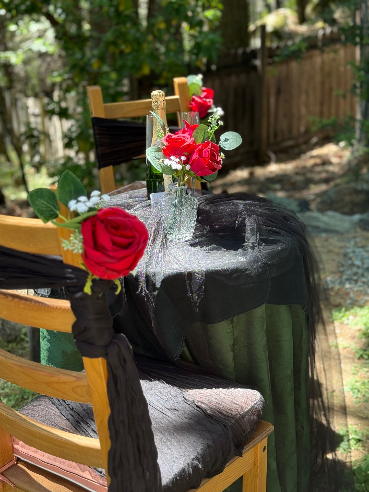 Red rose aisle chair flower rentals with black fabric ties on cross-back chairs at outdoor wedding table with black draping, champagne bottle, and red rose centerpiece | Reno, Lake Tahoe & Sacramento