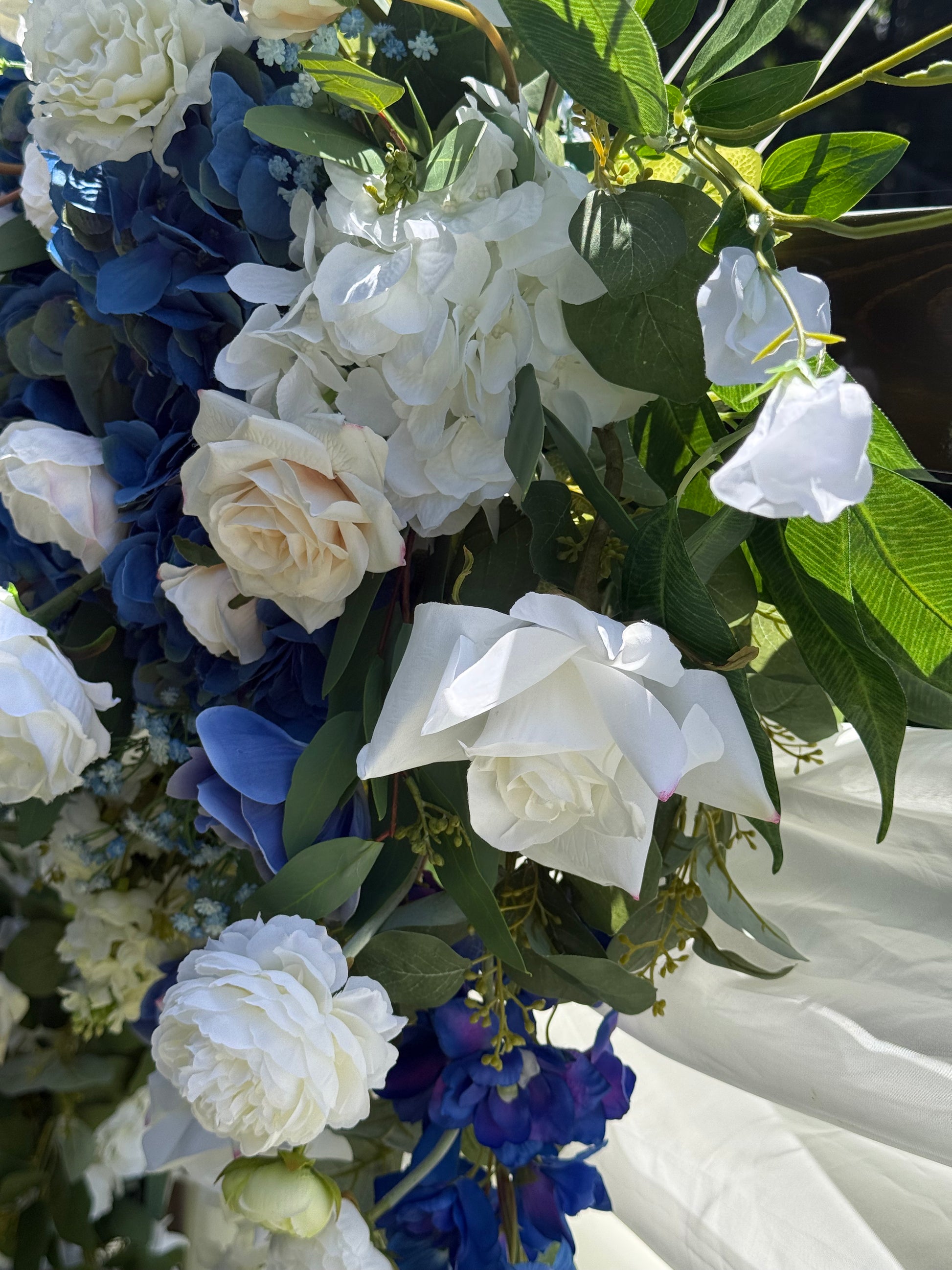 Close-up of Sweet Nothings arch adornment rental with white garden roses, blue hydrangea, and navy delphinium with white drapery in bright sunlight | Reno, Lake Tahoe & Sacramento