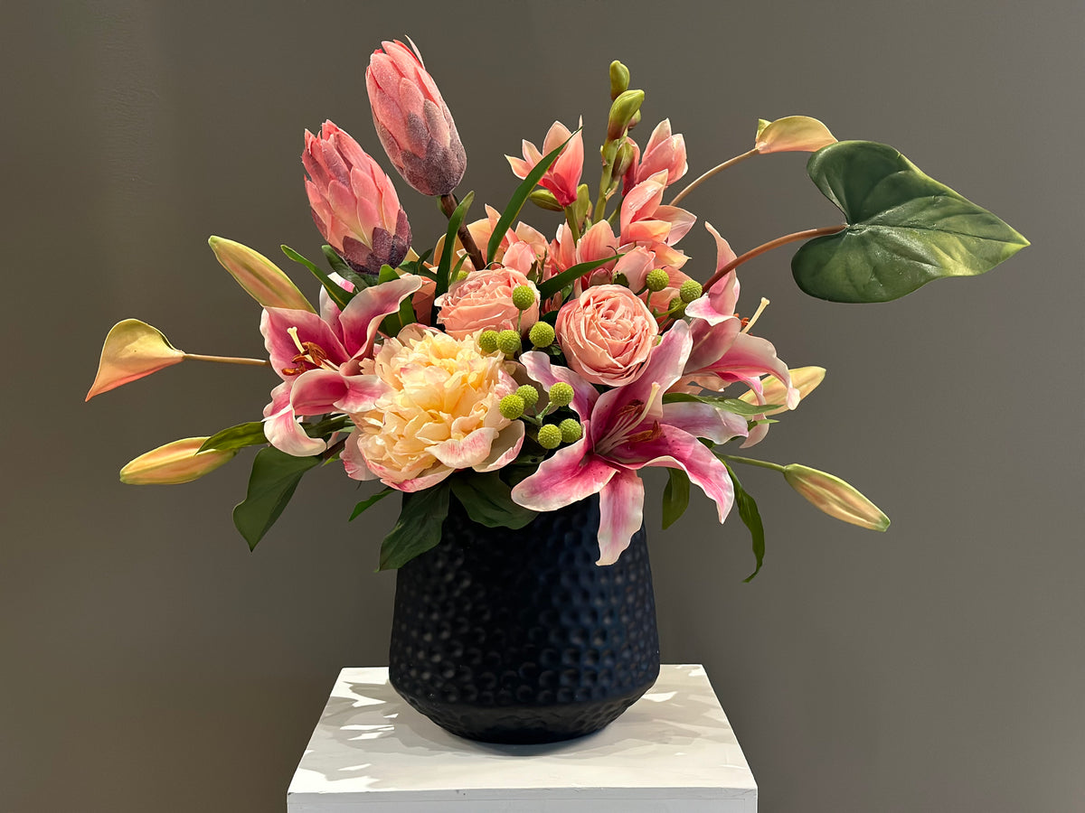 Tropical Blush centerpiece rental with pink stargazer lilies, blush protea, peach garden roses, blush peonies, hypericum berries, and tropical leaves in textured black vase on white pedestal | Reno, Lake Tahoe & Sacramento