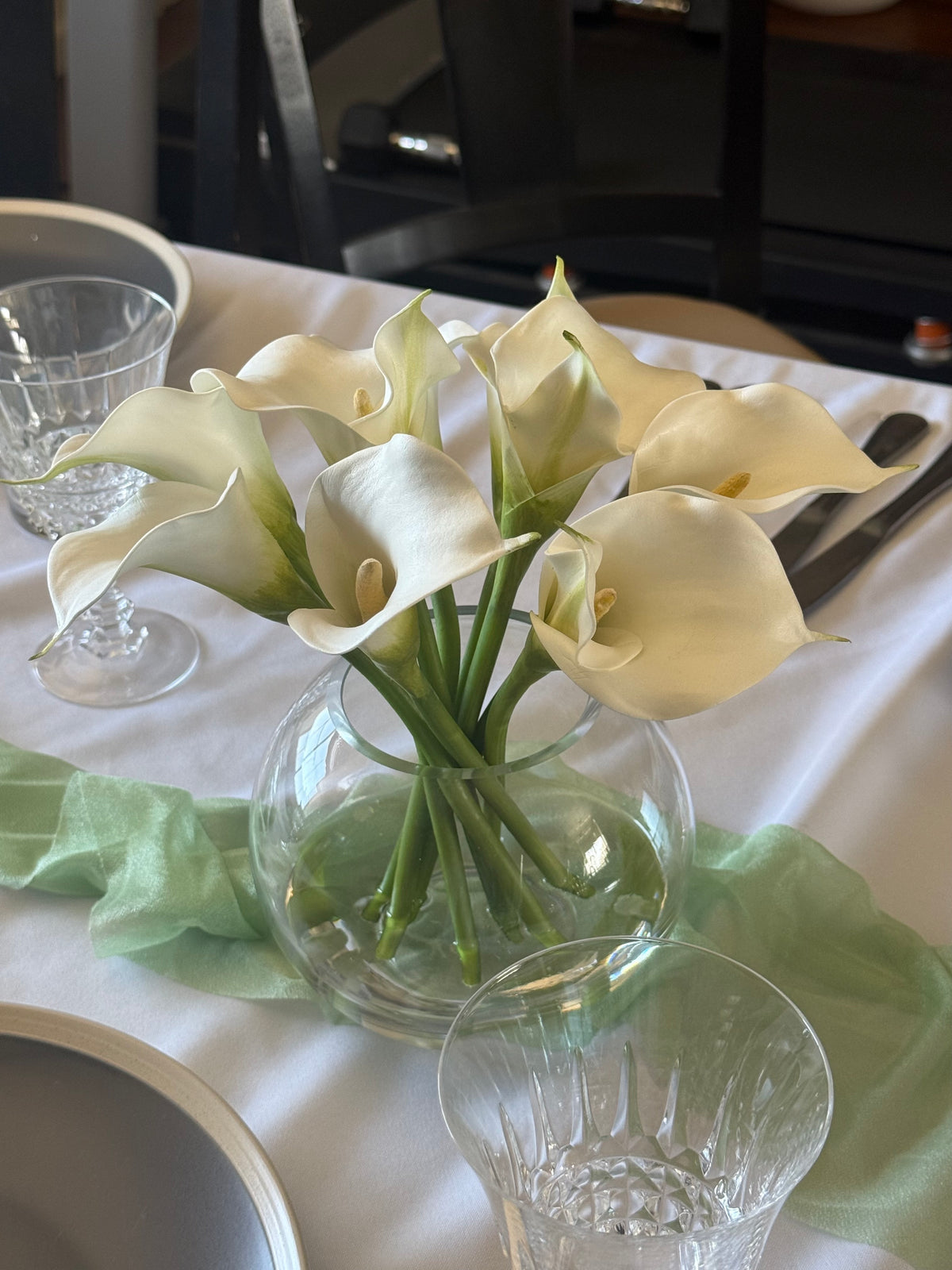 White Calla Lily centerpiece rental in round glass bubble vase on white linen wedding reception table with sage green napkins and crystal glassware | Reno, Lake Tahoe & Sacramento