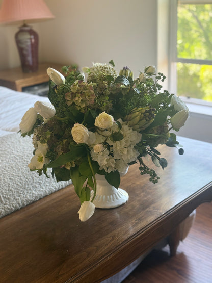 White Garden centerpiece rental with white tulips, white spray roses, white hydrangea, artichoke blooms, scabiosa, and lush greenery in white lattice compote vase on wood dresser at indoor home setting | Reno, Lake Tahoe & Sacramento