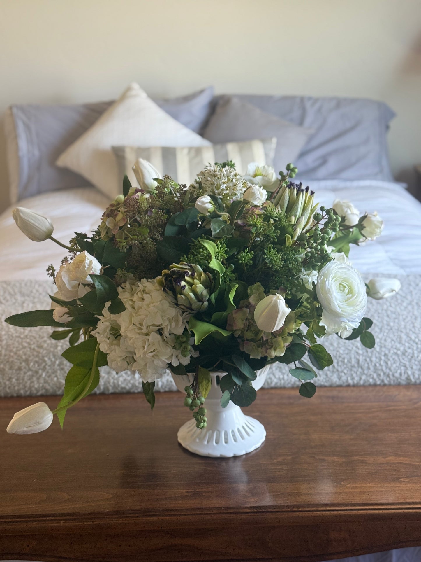 White Garden centerpiece rental with white ranunculus, white hydrangea, white tulips, artichoke blooms, scabiosa, and lush greenery in white lattice compote vase on wood side table at indoor home setting | Reno, Lake Tahoe & Sacramento