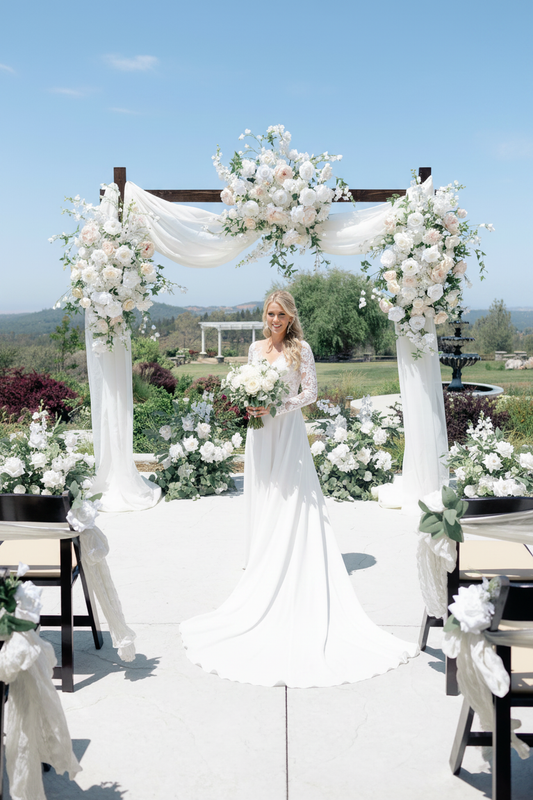 Bride walking down the aisle toward Love Story arch adornment rental with blush and white garden roses, delphinium, and white drapery on rustic wood arch at outdoor hilltop wedding venue | Reno, Lake Tahoe & Sacramento